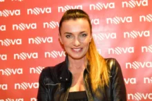 Žena stojí před plátněm s nápisy Nova, má vyčesaný koňský ohon a černou bundu. Usmívá se