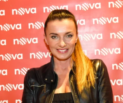 Žena stojí před plátněm s nápisy Nova, má vyčesaný koňský ohon a černou bundu. Usmívá se