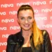 Žena stojí před plátněm s nápisy Nova, má vyčesaný koňský ohon a černou bundu. Usmívá se