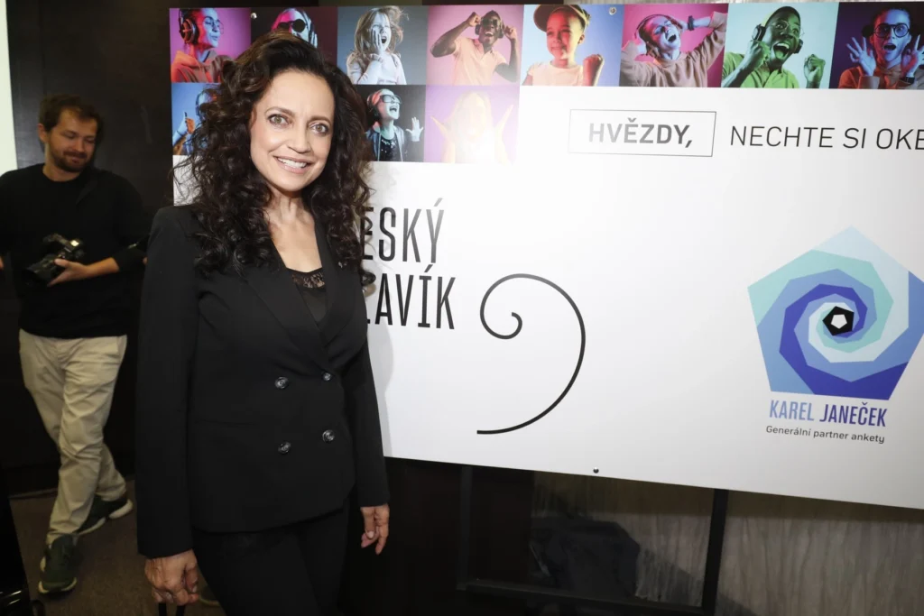 Žena stojí před tabulí s nápisy. Má černý outfit a kučeravé černé vlasy
