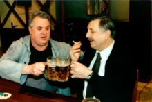 Dva muži si ťukají pivem v hospodě a jeden z nich kouří