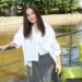 Smiling woman in a white blouse and gray pants leaning on a railing on a riverfront boat deck, with green chairs nearby and brown water in the background.