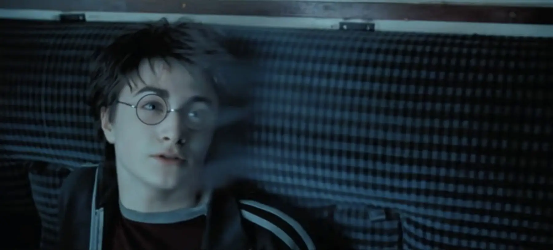 Person with round glasses and short dark hair lying down, looking off to the side against a blue striped background, nose ring visible.