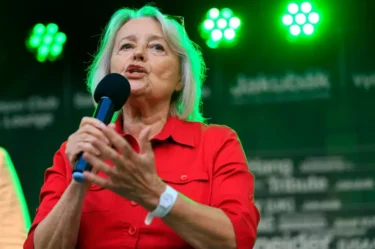 Žena v červené košili řeční na podiu a u úst si drží mikrofon