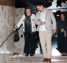Starší žena v krémovém kardiganu s černým lemováním sestupuje ze schodů zavěšená do mladého muže