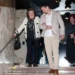 Starší žena v krémovém kardiganu s černým lemováním sestupuje ze schodů zavěšená do mladého muže