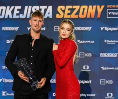 Muž v černém obleku drží trofej, o něj se opírá žena v rudých šatech