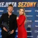 Muž v černém obleku drží trofej, o něj se opírá žena v rudých šatech