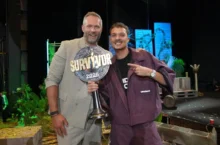 Two men smile on a stage, one holding a large 'SURVIVOR 2026' trophy between them.
