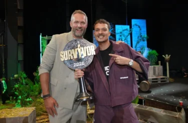 Two men smile on a stage, one holding a large 'SURVIVOR 2026' trophy between them.