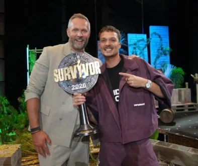 Two men smile on a stage, one holding a large 'SURVIVOR 2026' trophy between them.