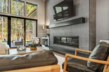 Modern living room with a built-in horizontal fireplace and textured gray tile wall, facing large glass doors to an outdoor seating area.