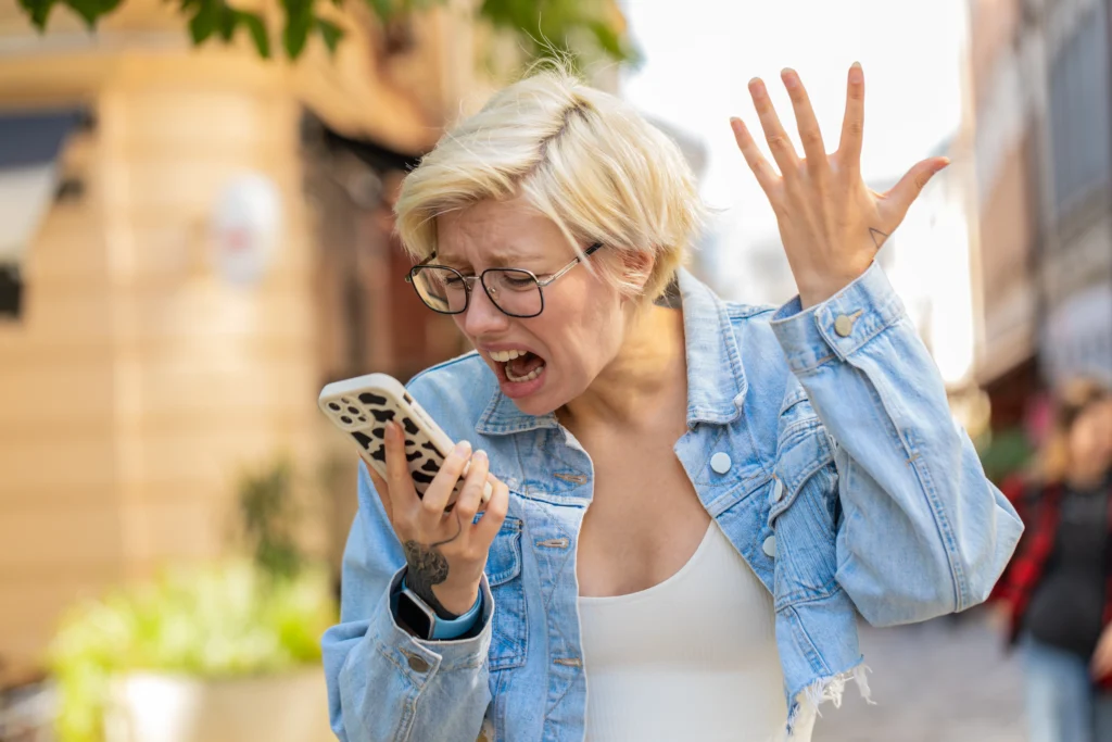shutterstock_2543906823 - VIPshow Žena na ulici v bílém tílku a modré košili nadává a gestikuluje do mobilního telefonu