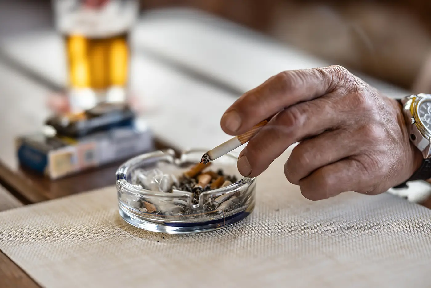 shutterstock_2648728275 - VIPshow Mužská ruka odklepává popel z cigarety do popelníku a nedaleko leží balíček cigaret a sklenice s pivem