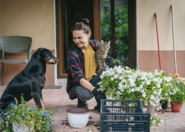 Žena sedí na verandě, v ruce drží kočku a usmívá se na svého psa.
