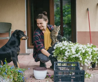 Žena sedí na verandě, v ruce drží kočku a usmívá se na svého psa.