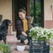 Žena sedí na verandě, v ruce drží kočku a usmívá se na svého psa.