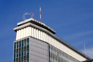 Logo společnosti České televize na rohu vysoké budovy, nad ní jsou modrá oblaka