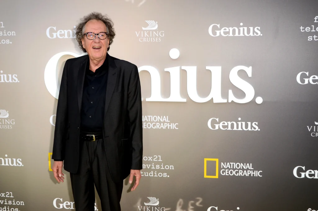 Muž v černém obleku a černé košili, na tváři má brýle, jeho vlasy jsou prošedivělé. Za ním je pozadí s nápisy Genius