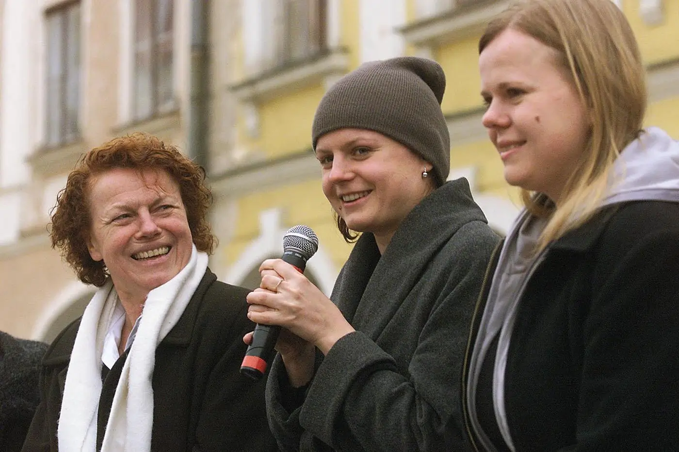 Three women outdoors; the middle woman speaks into a microphone while the others smile nearby.