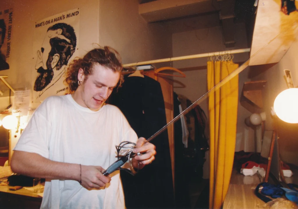 profimedia-0136919341 - VIPshow Young man in a white t-shirt using a soldering iron or tools while standing in a cluttered room with yellow curtains and a poster on the wall behind him.