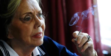 Stará žena v modrém saku a bílé košili kouří cigaretu