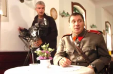Muž ve starodávné četnické uniformě sedí u stolku s bílým ubrusem, za ním je kameraman