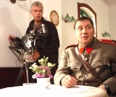 Muž ve starodávné četnické uniformě sedí u stolku s bílým ubrusem, za ním je kameraman