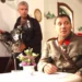 Muž ve starodávné četnické uniformě sedí u stolku s bílým ubrusem, za ním je kameraman