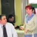 Zdravotní sestra s červenými vlasy hovoří s doktorem, on sedí, ona stojí a gestikuluje