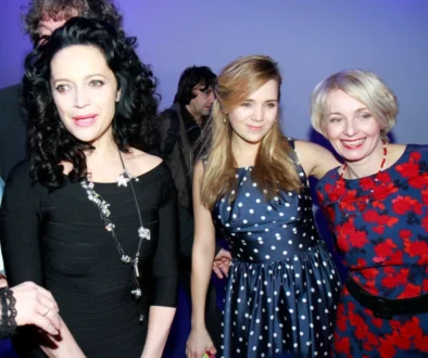 Tři ženy v letních slavnostních šatech na party, za nimi je modré pozadí a další lidé