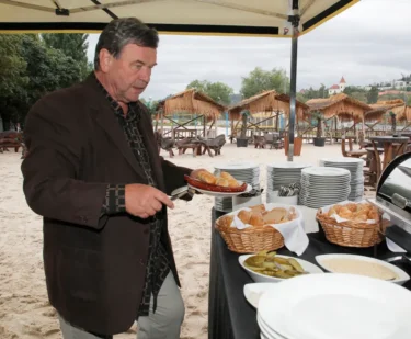 Muž v černém obleku stojí na písku a nabírá si jídlo ze stolu