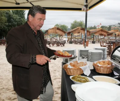 Muž v černém obleku stojí na písku a nabírá si jídlo ze stolu