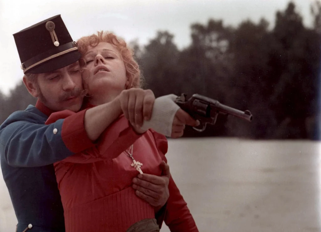 Žena v červených šatech a muž v historické uniformě míří starou pistolí, drží ženu