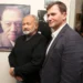 Dva muži v oblecích stojí u fotografie dalšího muže, synové a otec