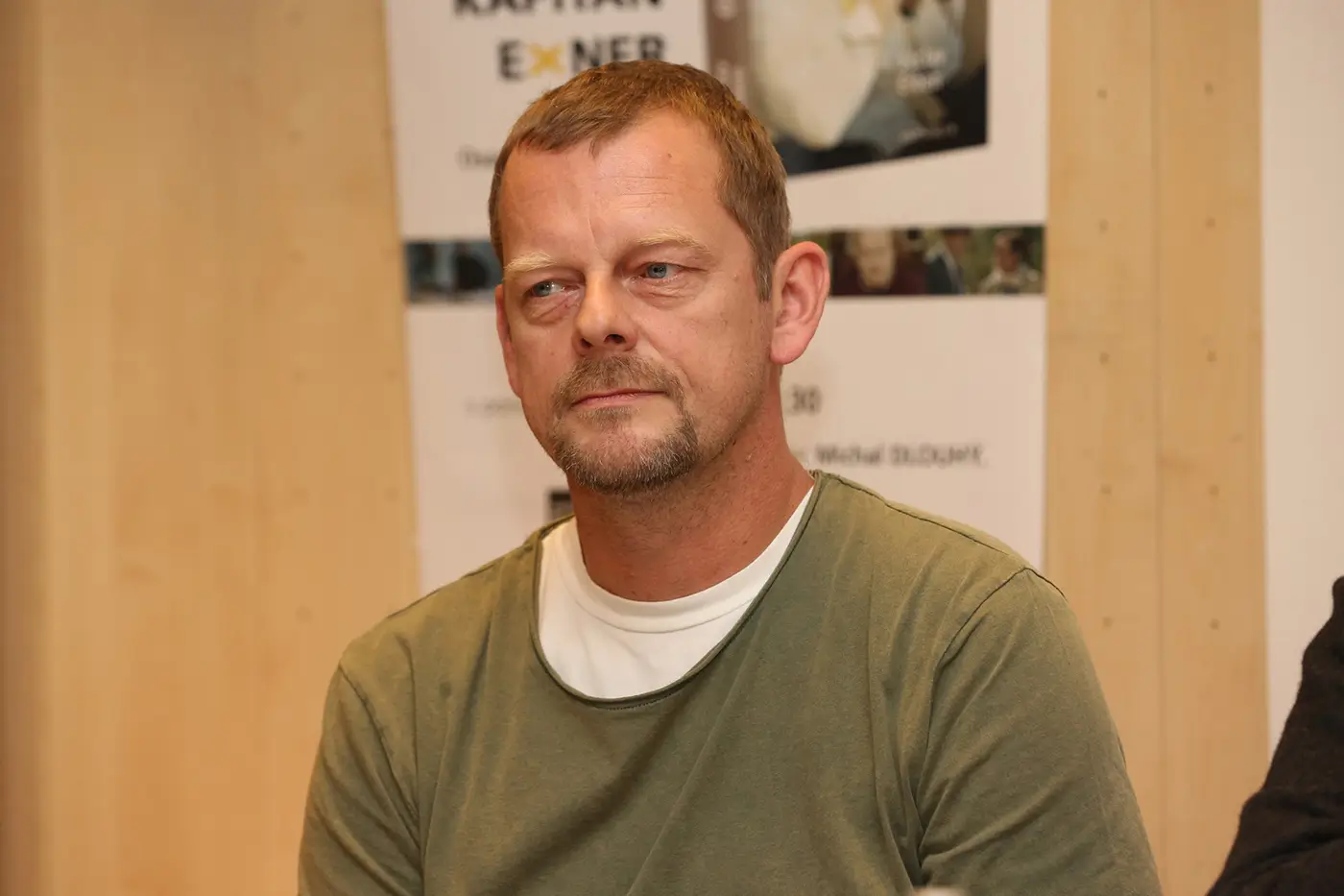 Muž v tričku a svetru se nechává fotit před plakátem a stěnou