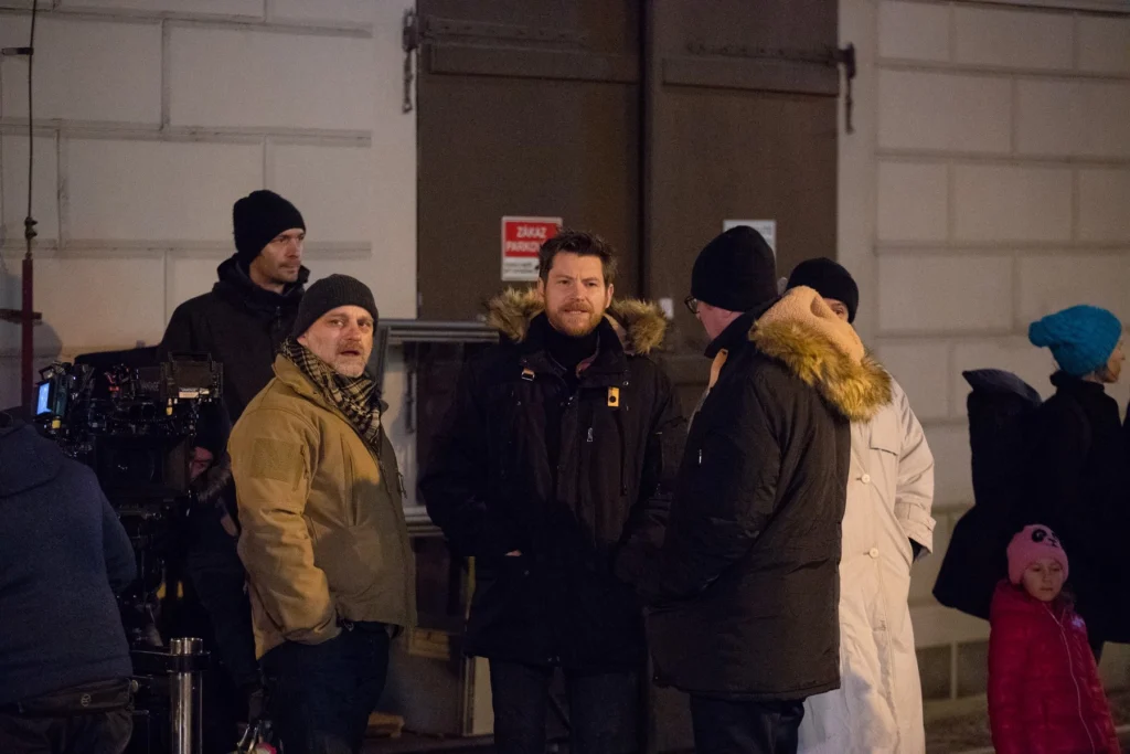 profimedia-0486712649 - VIPshow Group of people in winter coats and beanies standing together on a film set at night, with a camera rig visible on the left.