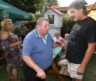 Dva muži si spolupovídají na zahradě u slunečníku, jeden je obézní, druhý vysoký