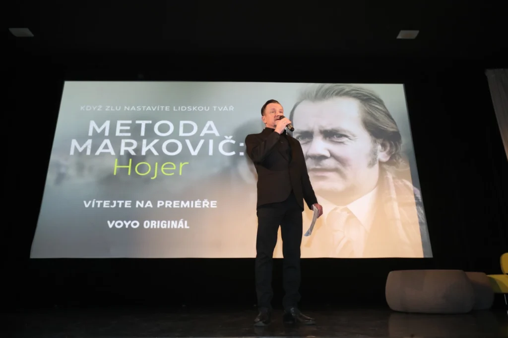 Muž v černém oblečení mluví do mikrofonu před filmovým plátnem, na něm je plakát seriálu Metoda Markovič: Hojer