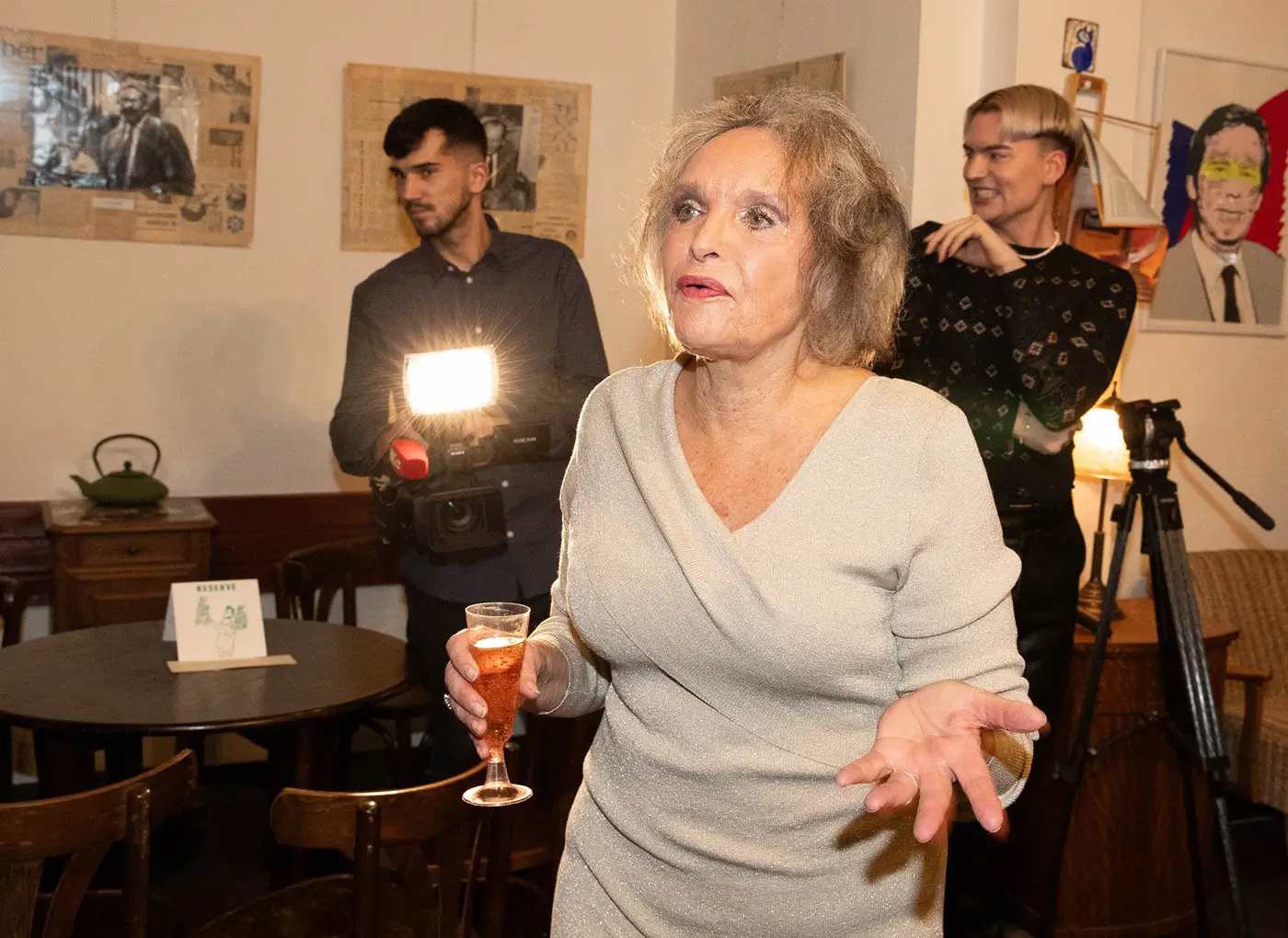 Starší žena ve světlých šatech gestikuluje a v pravé ruce drží skleničku