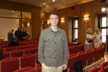 Young man in a gray patterned shirt standing in an auditorium with red seats and wooden walls in the background.