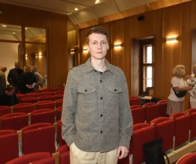 Young man in a gray patterned shirt standing in an auditorium with red seats and wooden walls in the background.