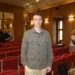 Young man in a gray patterned shirt standing in an auditorium with red seats and wooden walls in the background.