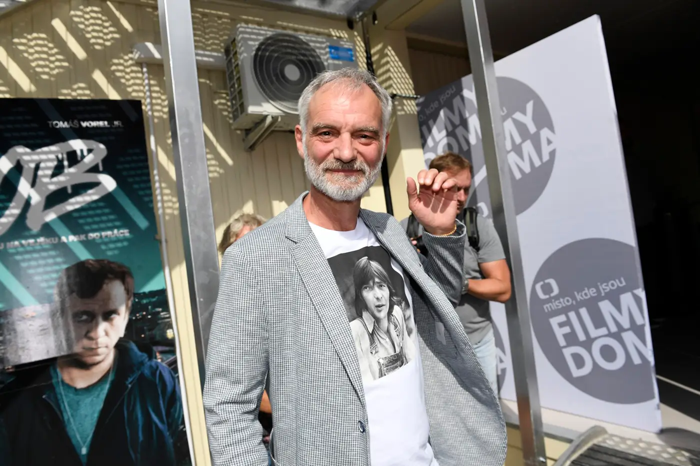 Muž v bílém tričku s fotkou a šedivém saku se usmívá