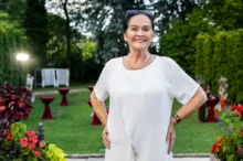 Smiling elderly woman in a white dress posing in a garden party setting with red cocktail tables and flowers nearby.