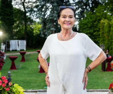 Smiling elderly woman in a white dress posing in a garden party setting with red cocktail tables and flowers nearby.