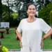 Smiling elderly woman in a white dress posing in a garden party setting with red cocktail tables and flowers nearby.