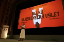 Žena v šatech stojí na podiu před plátnem promítajícím film