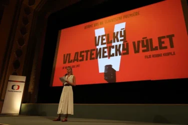 Žena v šatech stojí na podiu před plátnem promítajícím film
