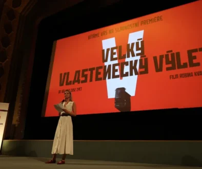 Žena v šatech stojí na podiu před plátnem promítajícím film